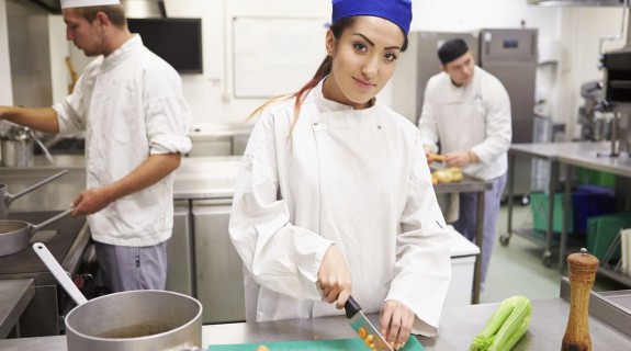A person in a purple cap and chef uniform cuts vegetables on a board in a kitchen